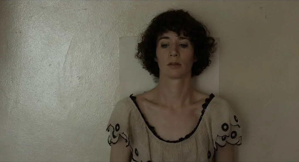 The Future (2011) movie still: medium — A woman stands against a blank, plain, off-white wall. She faces forward with a neutral expression while…