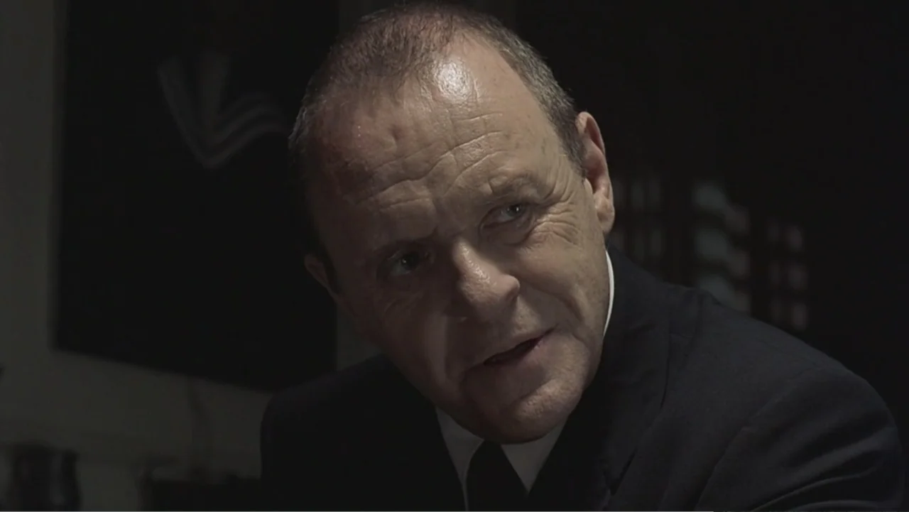 Hannibal (2001) movie still: closeup — A dark interior setting with a blurred wall element visible in the background. An elderly man in a dark suit…