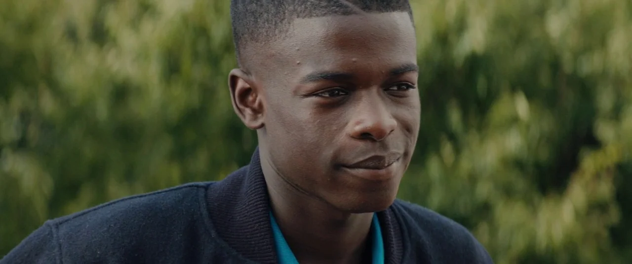 Girlhood (2014) movie still: closeup — A man stands outdoors with a background of blurred green foliage. The man looks off-camera with a neutral…