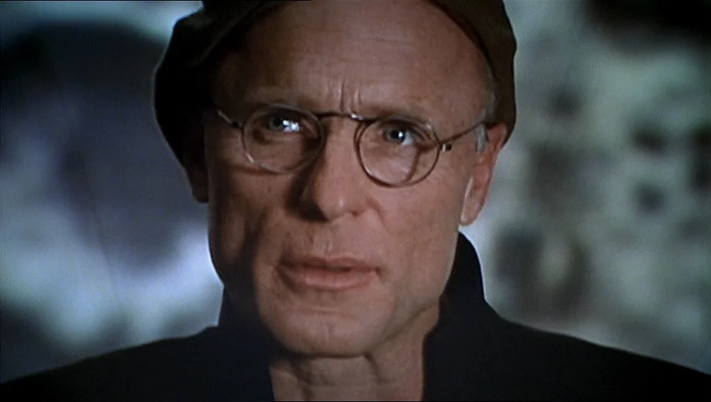 The Truman Show movie still: closeup — A dimly lit room features a glowing screen visible behind the man's head. The man wearing round glasses faces…