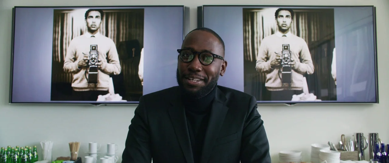 Yesterday (2019) movie still: medium — A modern meeting space features two large wall-mounted screens displaying the same photograph. A man in a…