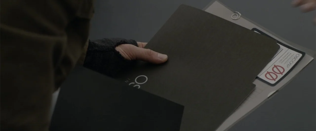 The Amazing Spider-Man (2012) movie still: closeup — A dark, smooth surface holds a black paper folder and a clear document sleeve. A hand in a…