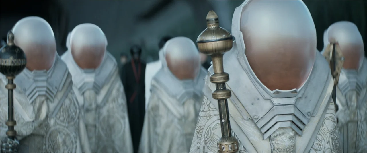 Dune (2021) movie still: medium — Several figures in white armored robes and domed helmets stand in an outdoor plaza. Each figure holds a tall…
