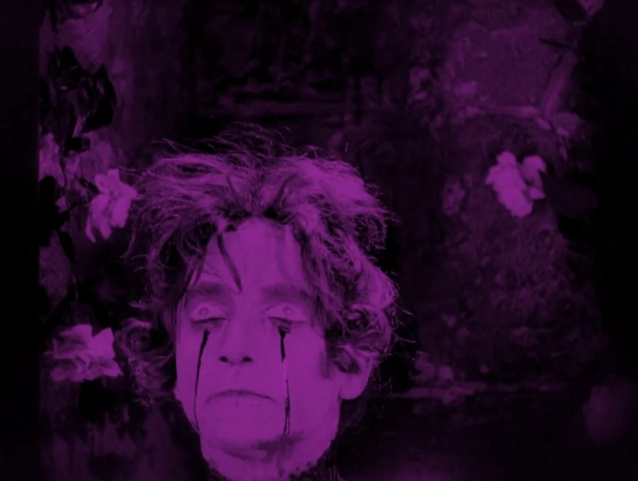 Napoleon (1927) movie still: closeup — A man's face appears centered against a dark, textured background with blurred flowers. Dark streaks…