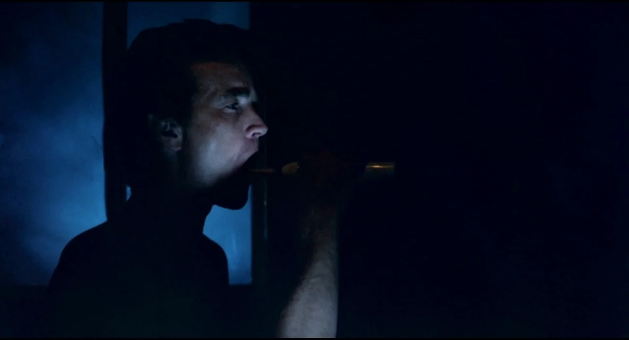 Olivia (1983) movie still: medium — A dark, dimly lit space is dominated by cool blue light from a single source. A man is captured in profile while…