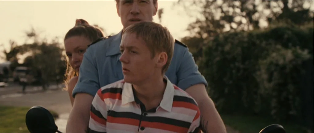 The Scouting Book for Boys (2009) movie still: medium — Two men ride a moped along an outdoor road at golden hour. The sunlight casts soft…