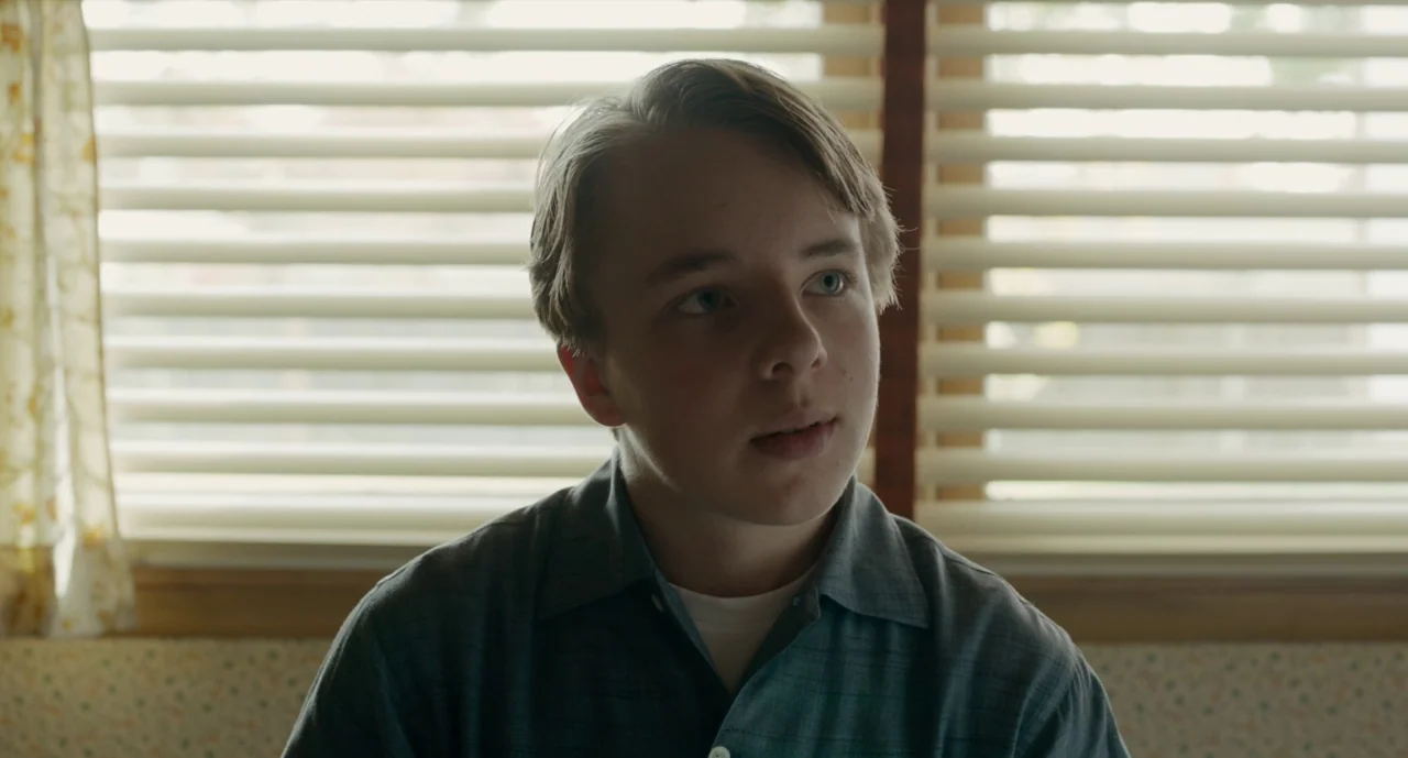 Wildlife (2018) movie still: medium — An interior space with horizontal window blinds and patterned walls. A teenage boy sits facing the camera…