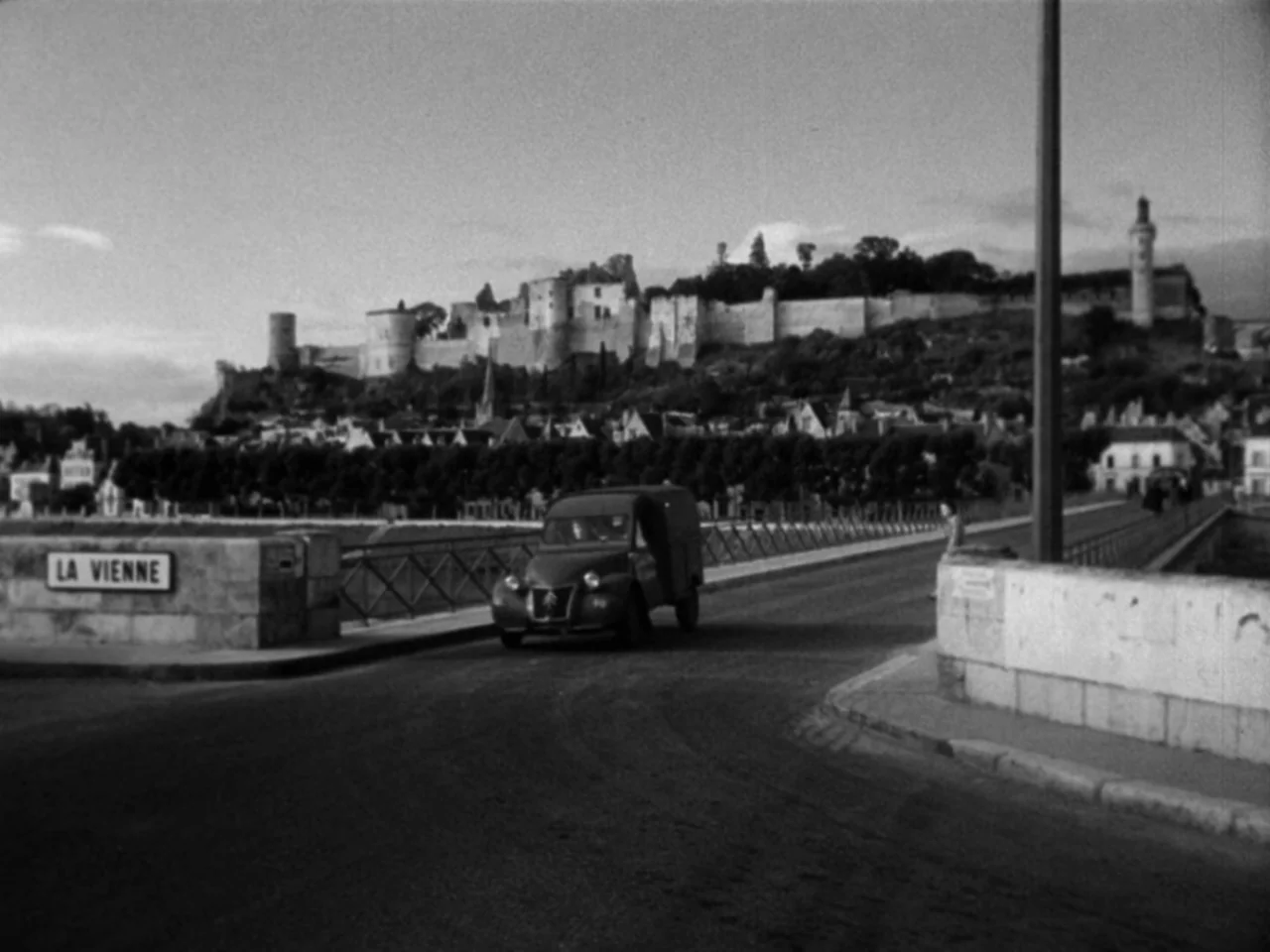 Diabolique (1955) movie still: establishing — An old stone bridge crosses the river Vienne toward a walled medieval town hillside. A small dark van…