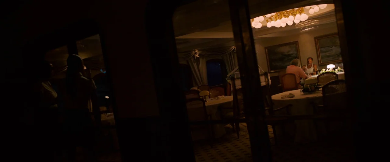 Triangle of Sadness (2022) movie still: wide — A dimly lit, formal dining hall features round tables and hanging chandeliers at night. Two men are…