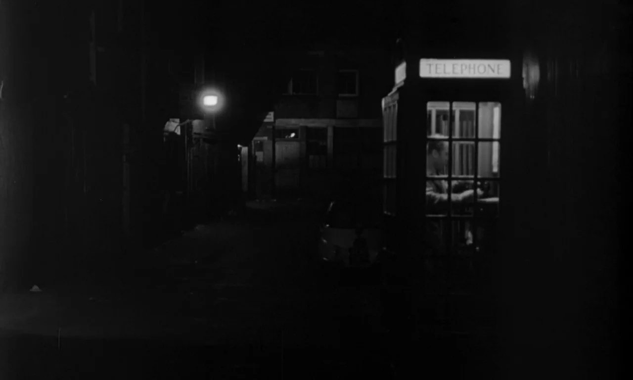 Seance on a Wet Afternoon (1964) movie still: wide — A dark, empty urban street at night with a single bright telephone box. A man is visible inside…