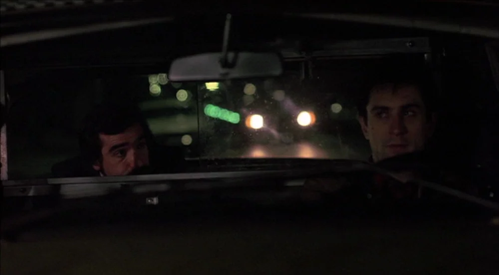 Taxi Driver (1976) movie still: medium — The interior of a taxi cabin at night is darkened with two men sitting forward. Headlights from the…
