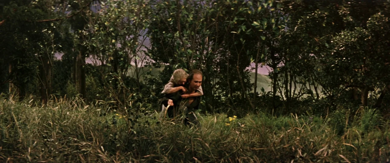 Ballad Of Narayama (1958) movie still: wide — A dense patch of tall wild grass and thin trees fills the frame. A man carries an elderly woman on his…