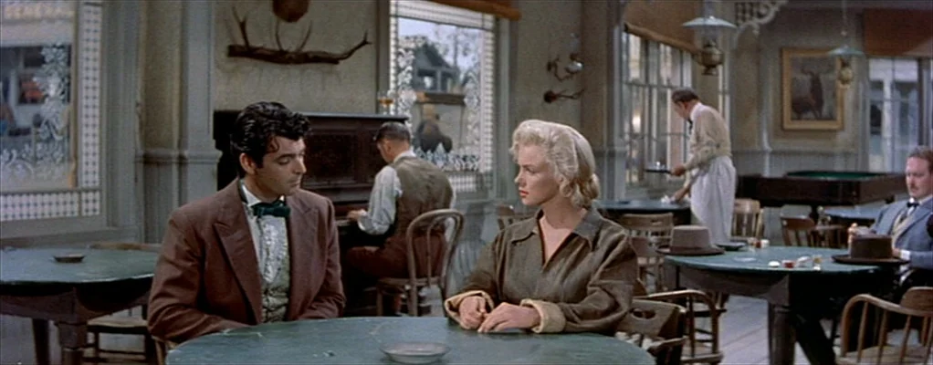 River of No Return (1954) movie still: medium — A 1940s western saloon interior features green round tables and wood-paneled walls. A man and woman…