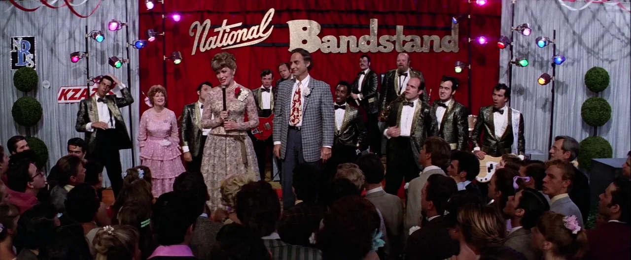 Grease (1978) movie still: wide — A televised bandstand set features a large red curtain backdrop with gold lettering. A middle-aged man and woman…