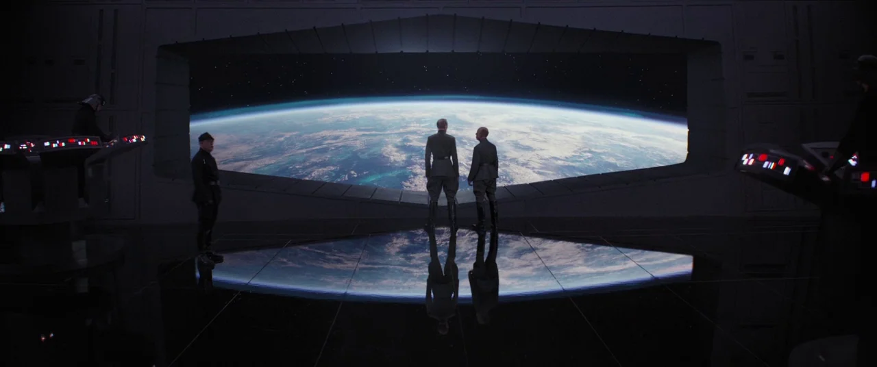 Rogue One: A Star Wars Story (2016) movie still: wide — A dark, expansive room features a wide viewport looking out at a curved planet. Two officers…