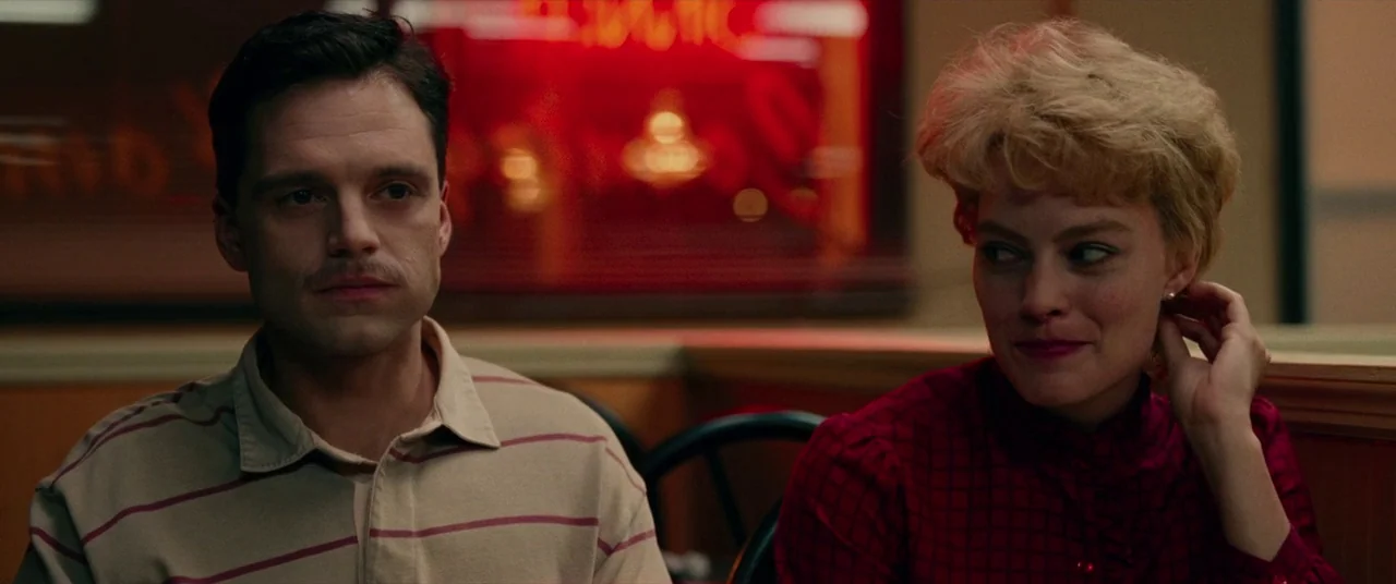 I, Tonya (2017) movie still: medium — A diner booth with wooden partitions and a glowing red neon sign in back. A man and a woman sit together…