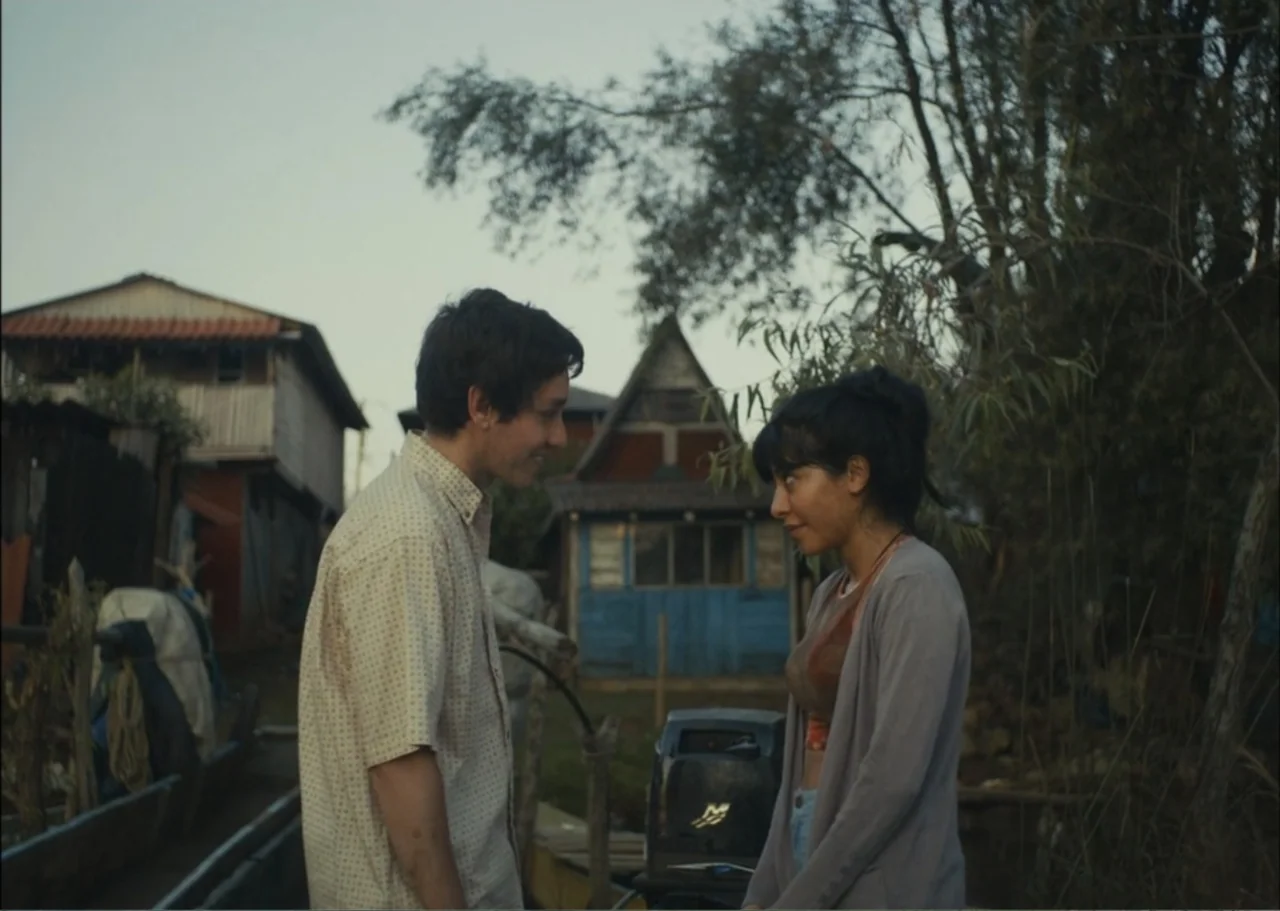 A Fisherman's Tale (1976) movie still: medium — A rural setting features simple wooden buildings and sparse vegetation under an overcast sky. A man…
