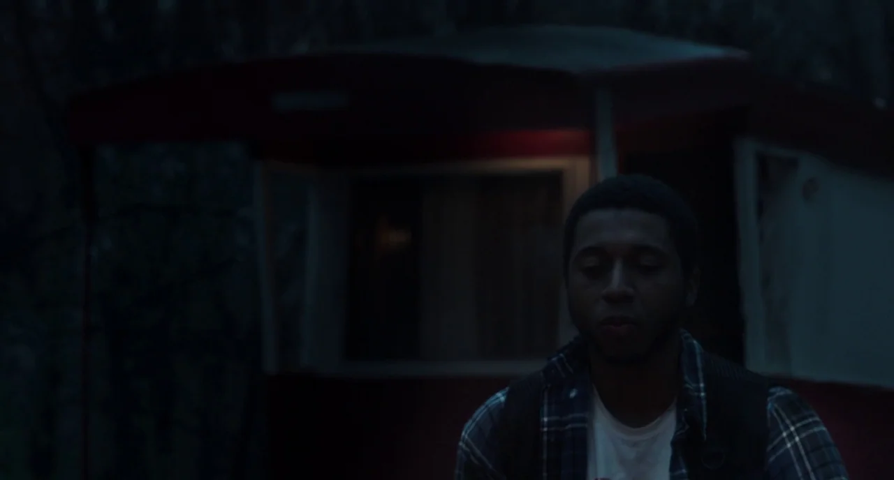 The Alchemist Cookbook (2016) movie still: medium — A man sits outdoors at night in front of a dark, stationary trailer home. The background is dim…