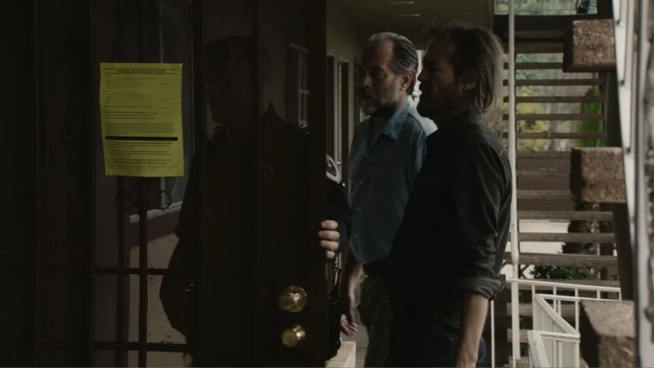 The Cleaner (2021) movie still: medium — Two men stand on the exterior walkway of an aging, multi-level apartment or motel complex. One man stands…