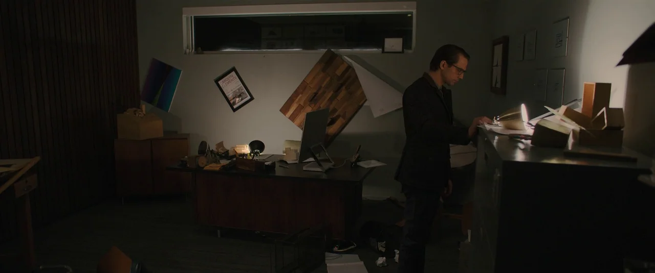 Intrusion (2021) movie still: medium — A dimly lit architectural studio features geometric wooden art and a cluttered dark desk. A man in a dark…