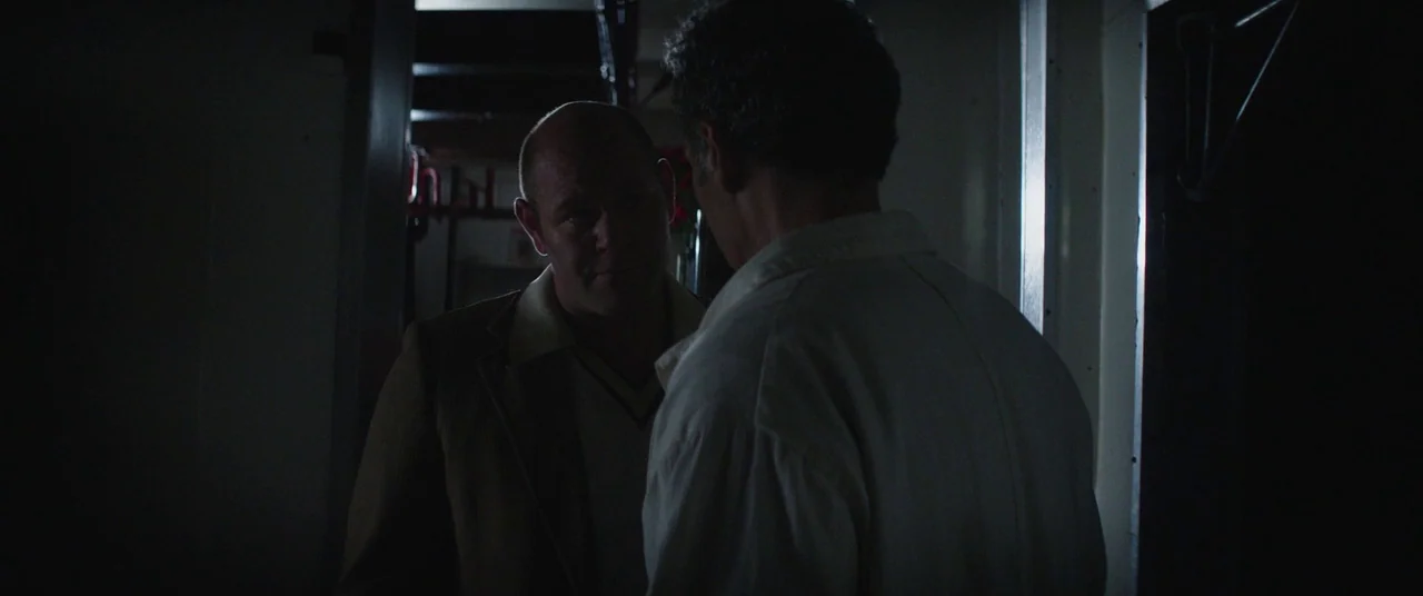 God’s Pocket (2014) movie still: medium — A dimly lit narrow hallway interior features dark walls and minimal structural details visible. Two men…