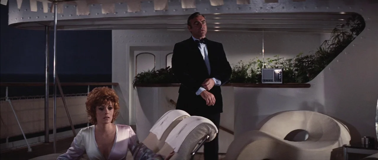 Diamonds Are Forever (1971) movie still: medium — The deck of a luxury yacht at night features white lounge chairs and plants. A man in a tuxedo…