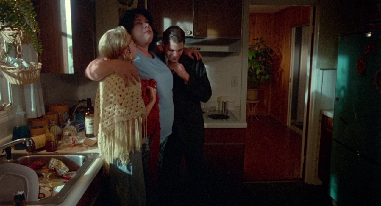 Polyester (1981) movie still: medium — A residential room featuring dark wooden cabinetry and a cluttered metal sink area. Three people are standing…