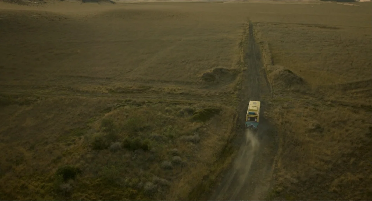 Ice Road: Vengeance (2025) movie still: establishing — A vast, dry grass field is bisected by a single narrow dirt track. A small yellow camper van…