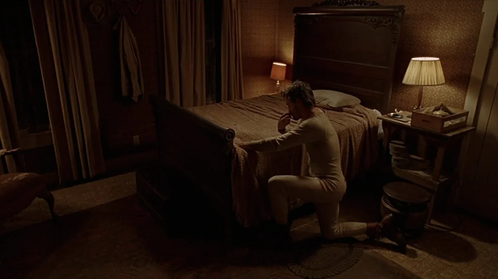 Undertow (2004) movie still: medium — A dimly lit 1940s bedroom has wallpaper, a dark wooden bed frame, and lamps. A man in thermal clothing kneels…