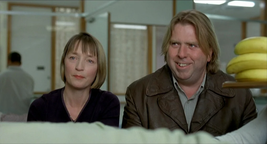 All or Nothing (2002) movie still: medium — A brightly lit interior space with glass partitions and wood-framed walls. A man and woman sit next to…