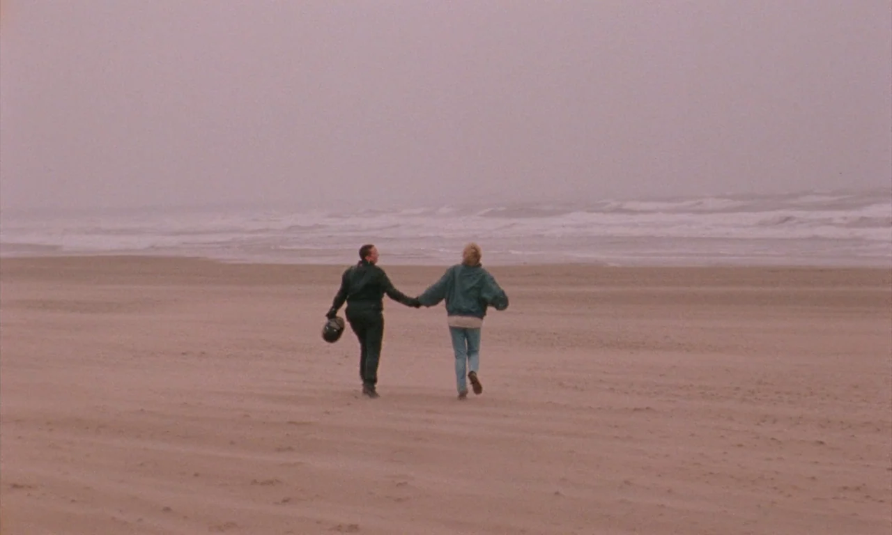 Blue Jean (2022) movie still: wide — A wide, flat, overcast beach with light sand and surf in the distance. A man holding a helmet and a woman run…