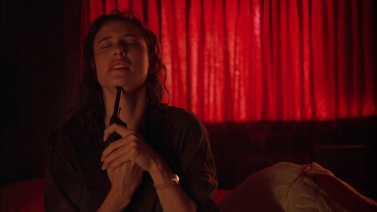 The Rapture (1991) movie still: medium — A dark bedroom with bright red curtains covering the window in the background. A woman sits on a bed…