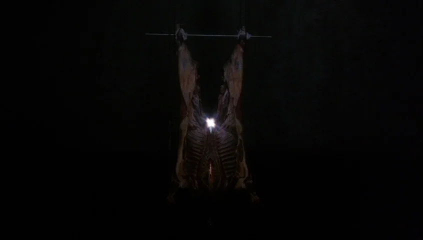 Edward II (1991) movie still: wide — A dark, empty space contains a single animal carcass suspended from a metal bar. The carcass is illuminated by…