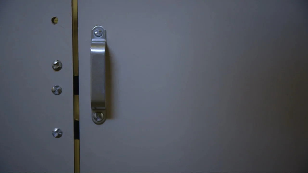 Beware the Slenderman (2016) movie still: closeup — A close-up view shows the metallic surface of a gray bathroom stall door. A simple silver handle…