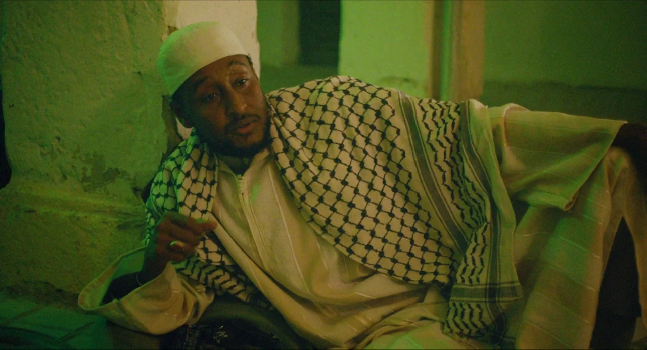 Submergence movie still: medium — A dimly lit room with textured, distressed walls and a green light source. A man wearing a white cap and checkered…
