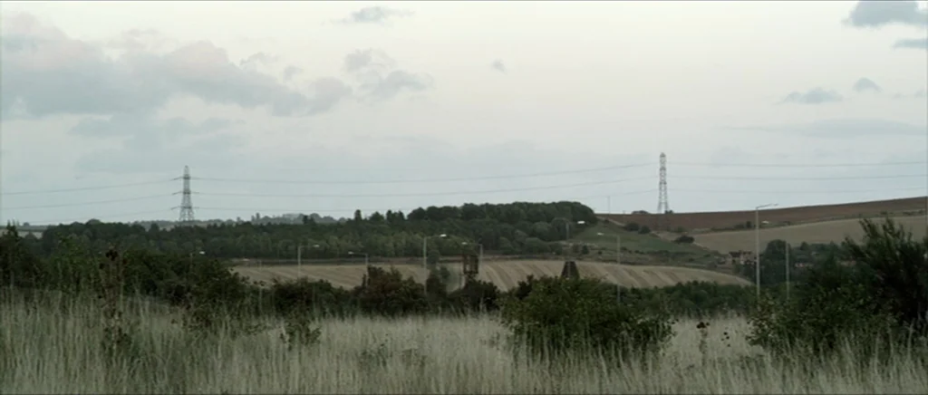 Kill List movie still: wide — A rural landscape with rolling fields, dense green treelines, and tall power pylons. Overcast sky hangs above the…