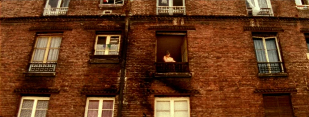 Carne (1991) movie still: establishing — The red brick exterior of a multi-story apartment building features repeating window patterns. A single…