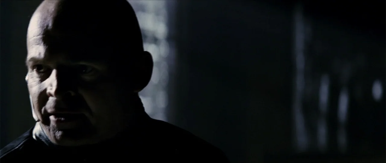 Elektra (2005) movie still: closeup — A dark indoor setting with minimal light illuminates only part of a man's face. The subject stares ahead in a…