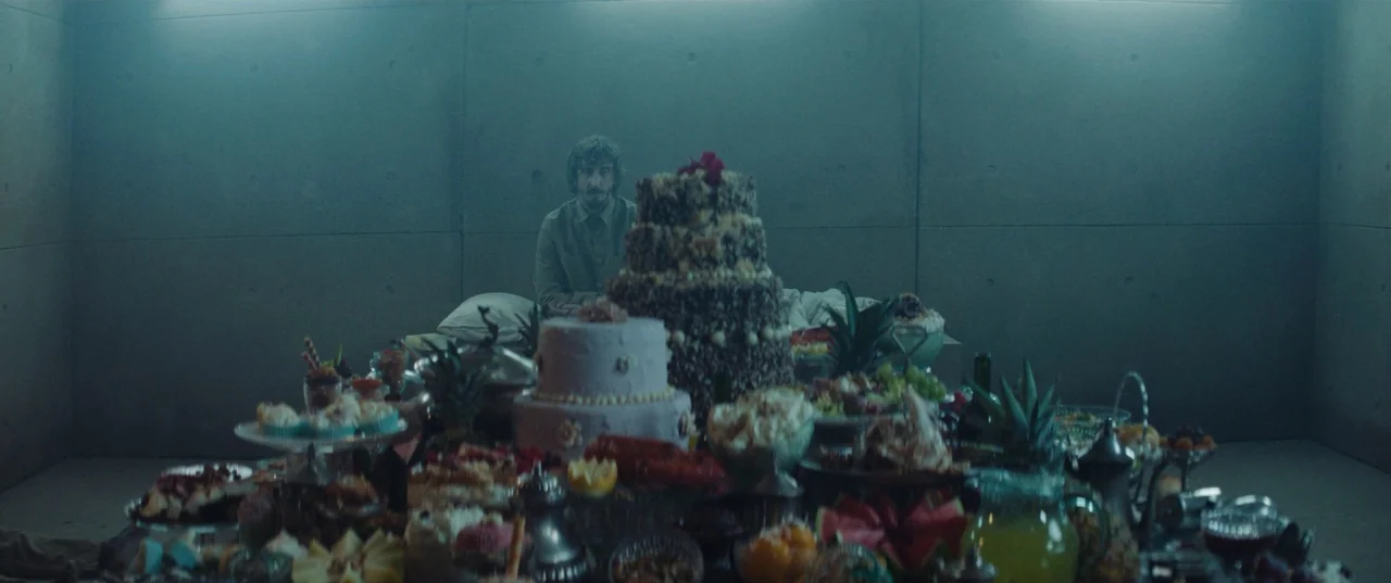 The Platform (2019) movie still: wide — A stark, concrete-walled room contains a bed and a table crowded with gourmet food. The man sits on the bed…