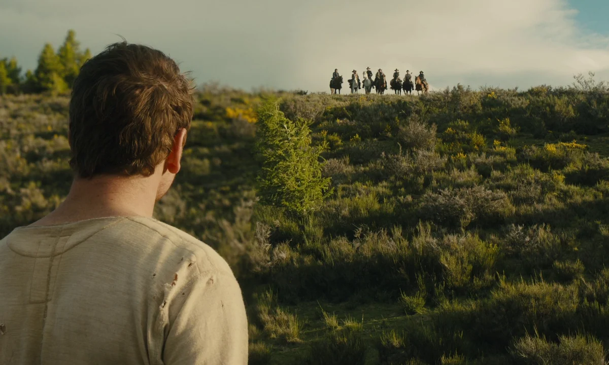Slow West (2015) movie still: medium — A man stands in a brush-covered landscape looking toward a distant grassy hill. A group of several riders on…