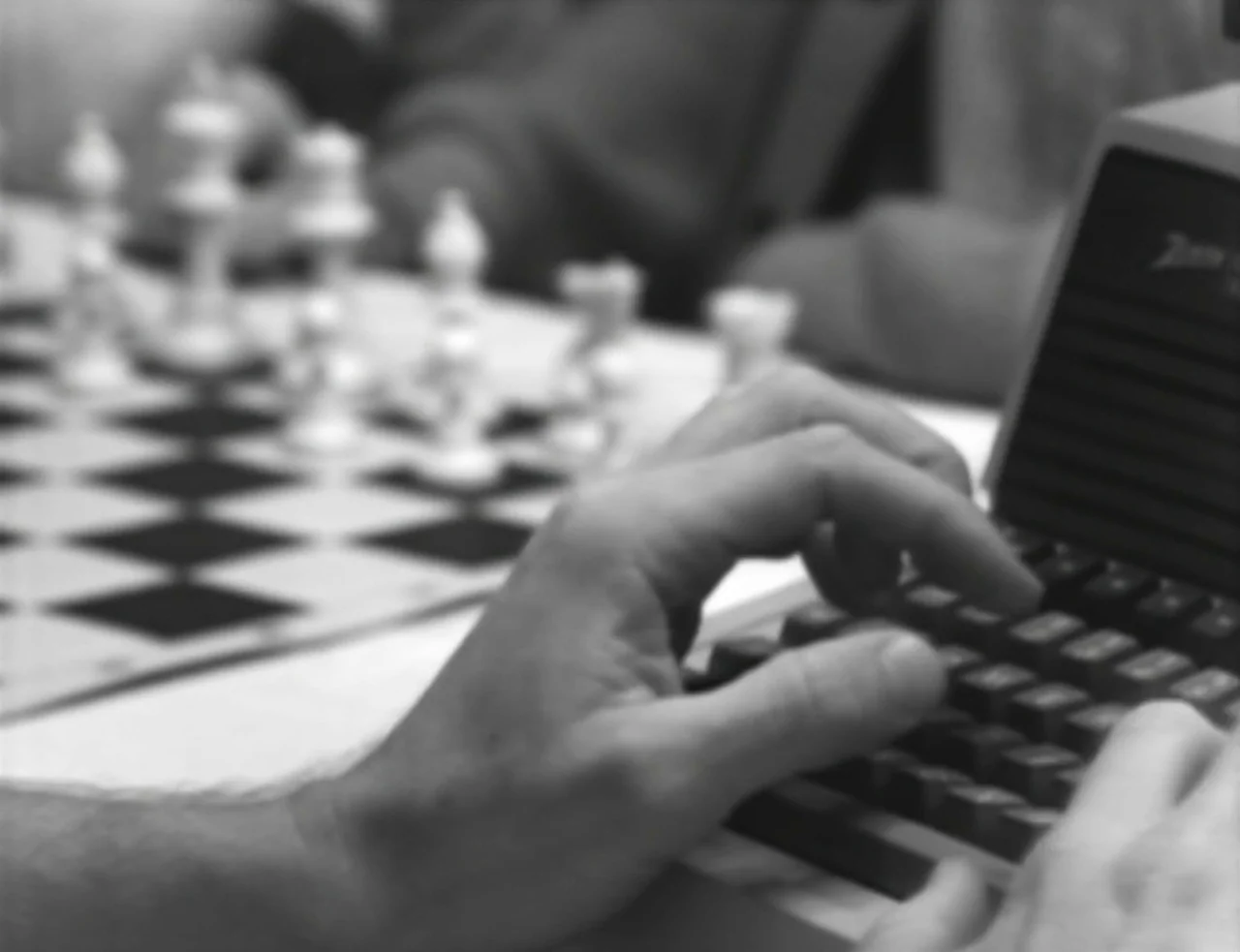 Computer Chess (2013) movie still: closeup — A pair of hands types on a portable electronic keyboard on a white table. A blurred chessboard and game…