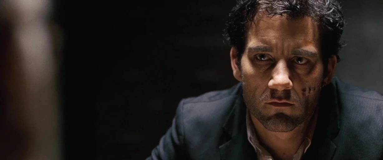 The International (2009) movie still: closeup — A dimly lit room with dark walls and minimal environmental detail visible. A man with facial…