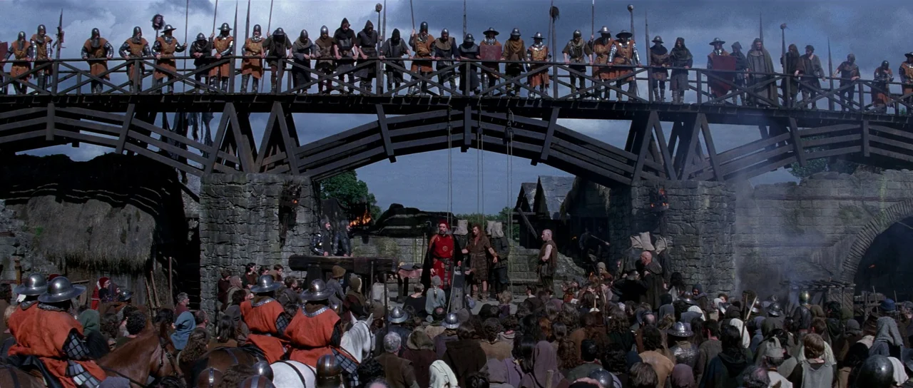 Braveheart (1995) movie still: wide — A wooden bridge spans a stone gateway structure within a medieval village setting. A large group of soldiers…