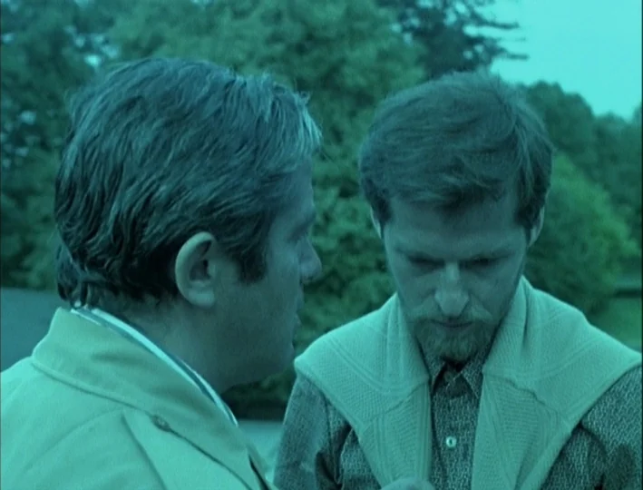 Camera Buff (1979) movie still: medium — Two men stand outdoors near blurred green trees under an overcast sky. One man listens to the other while…