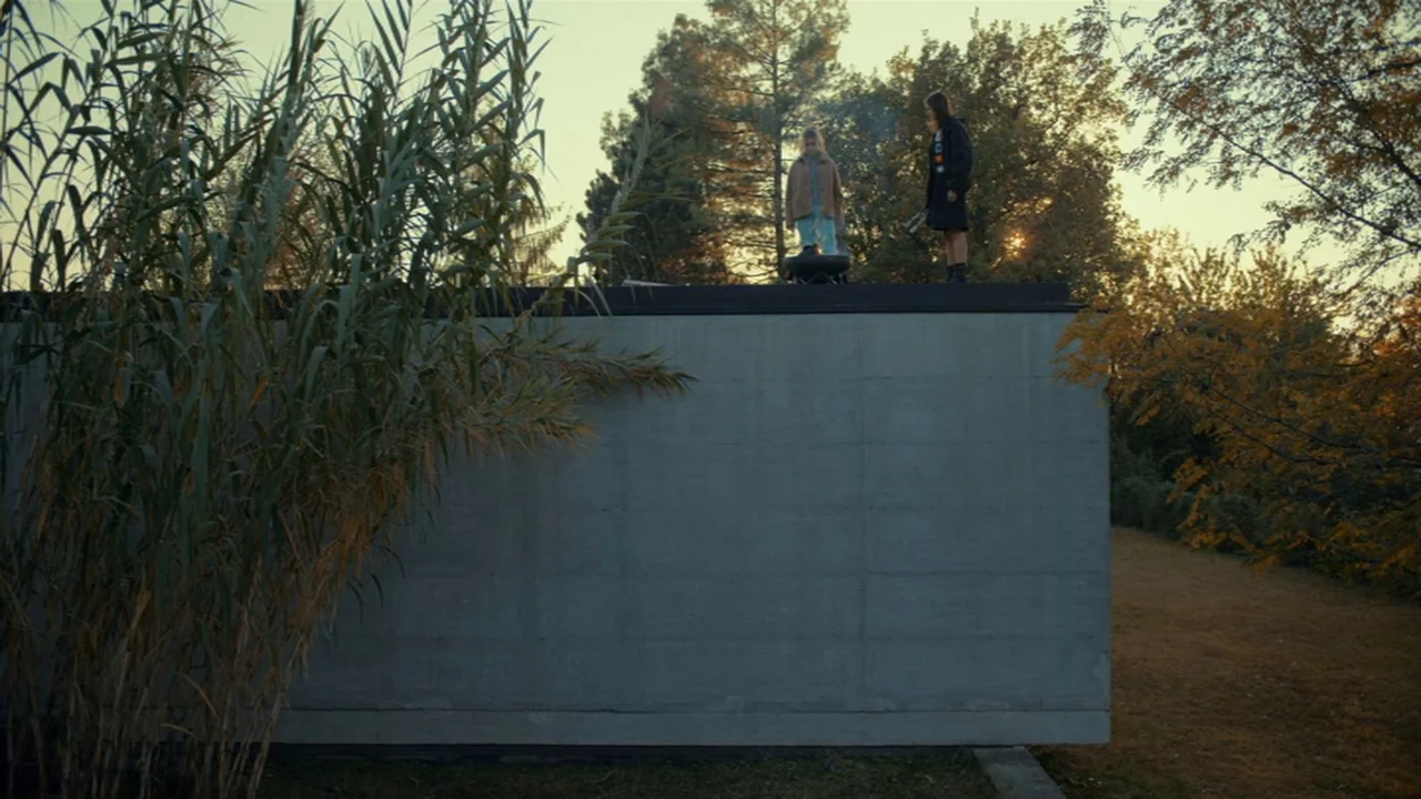 Amanda (2022) movie still: wide — Two individuals stand on the flat roof of a modern concrete building during sunset. They are gathered around a…