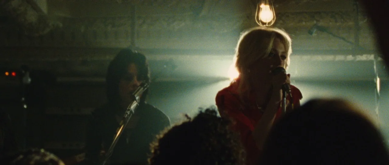 The Runaways (2010) movie still: medium — Two female musicians perform on a dimly lit stage in a crowded room. A blonde singer holds a microphone…
