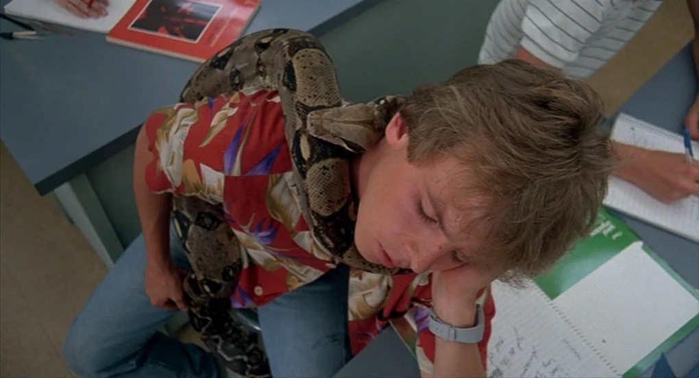 A Nightmare On Elm Street 2: Freddy’s Revenge (1985) movie still: medium — A student sits at a grey laminate desk with notebooks and a red textbook.…