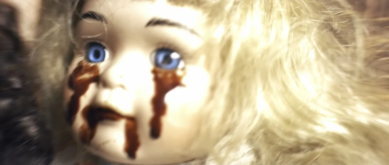 The Lullaby (2019) movie still: closeup — A porcelain doll with blonde hair and blue eyes rests in an undefined space. Dark liquid streaks run from…