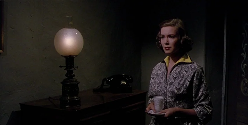 Magnificent Obsession (1954) movie still: medium — A dimly lit interior space with a dark wooden table against a textured wall. A woman in a…