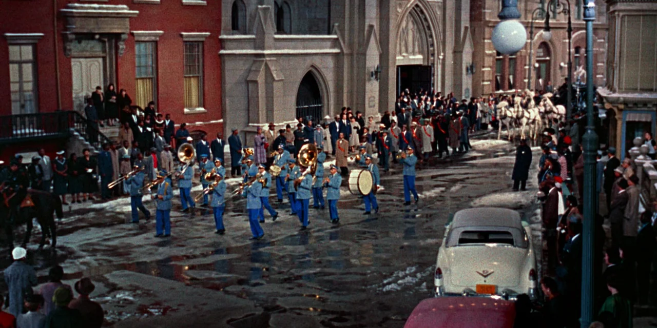 Imitation of Life (1959) movie still: wide — A city street with wet pavement and brick buildings hosts a public procession. A large marching band in…